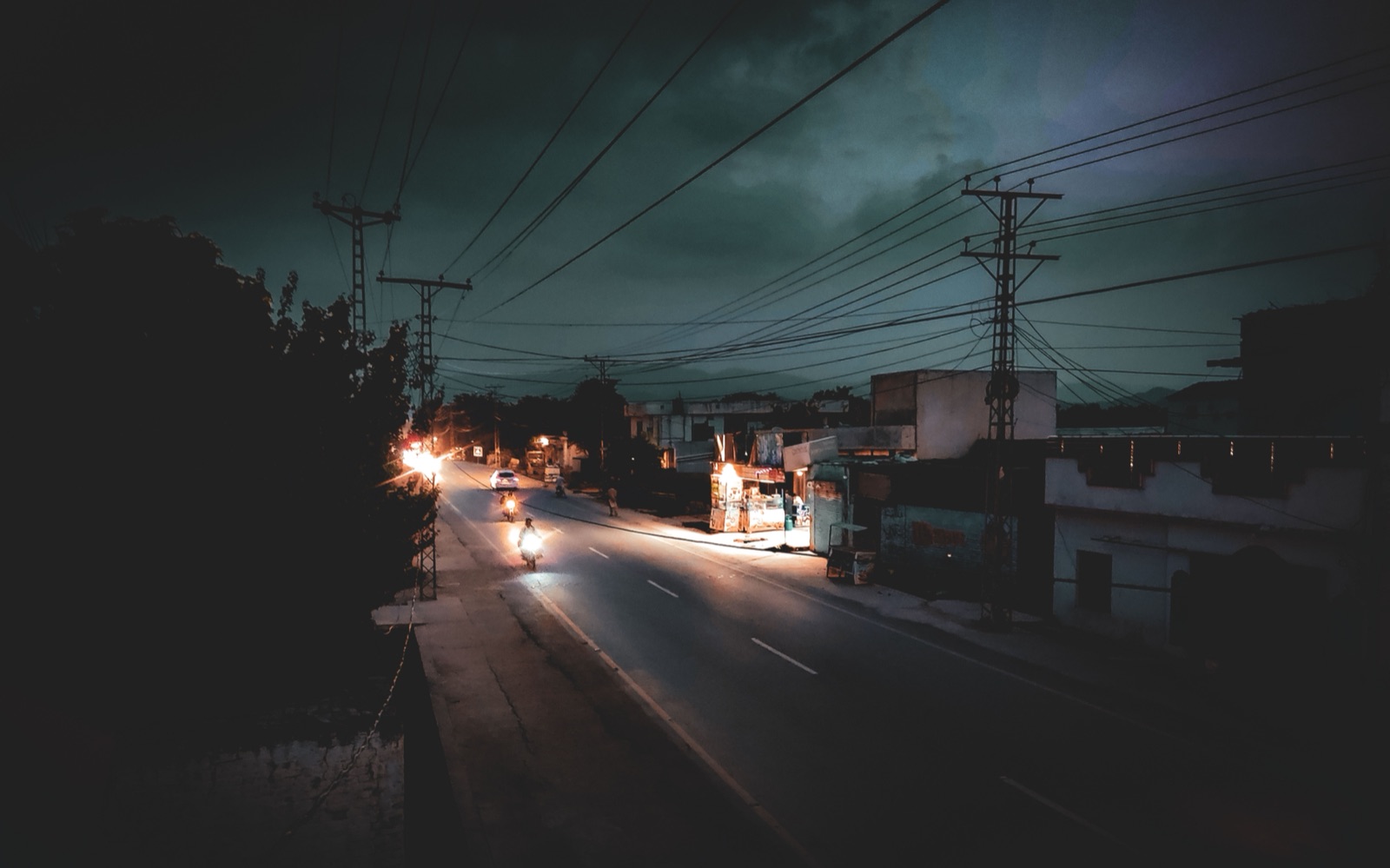 Calle urbana vacía en una noche lluviosa y oscura, con postes de energía y un resplandor lejano.